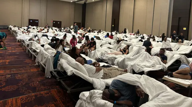 Dozens of people sit on cots with white sheets in a hotel ballroom