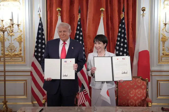 Tổng thống Hoa Kỳ Donald Trump và Thủ tướng Nhật Bản Sanae Takaichi, mỗi người giơ cao một tài liệu có chữ ký của họ với cờ Hoa Kỳ và Nhật Bản phía sau