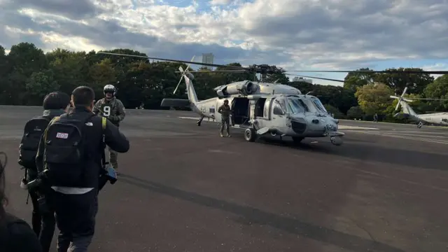 Shot of a helicopter on the tarmac, with guards and reporters walking towards it