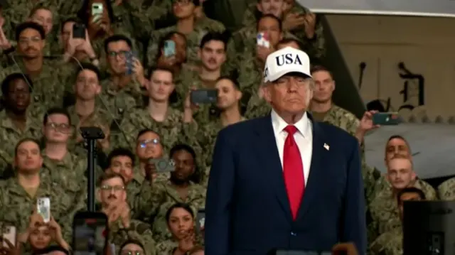 Trump wearing a USA cap wearing a suit, with military uniformed people behind him