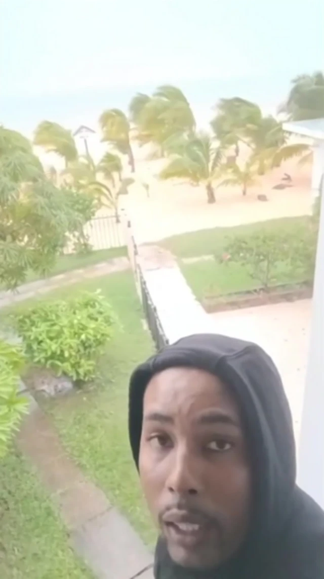 A still of Shaquille Clarke as he videos himself on his phone - trees can be seen bending behind him in the wind