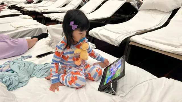 A little girl in pyjamas watches TV on her tablet while in a hotel shelter