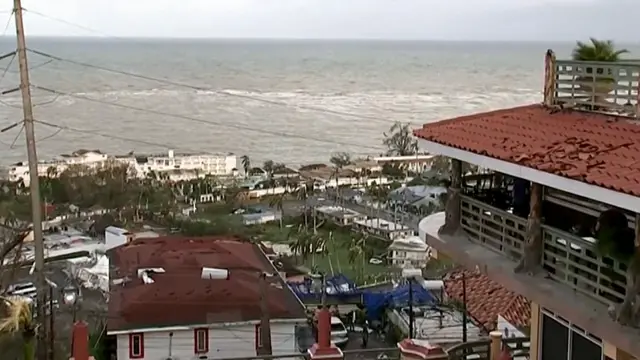 A still of damage to buildings near the coast in Jamaica, debris on the ground and trees blowing in the wind.