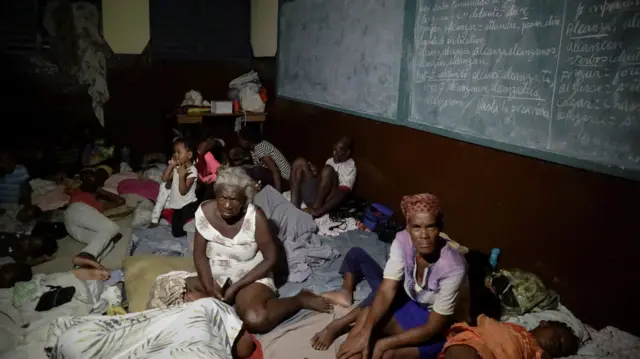 Adults and children sit on the floor beneath chalk boards. Some are asleep and some are sitting up, looking into the camera.