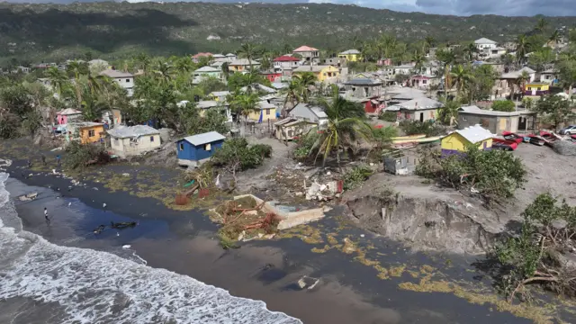 A beach is shown from the air with debris across it, trees uprooted and house with sand eroded from underneath