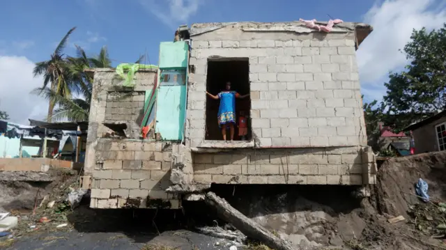 A woman looks out of the doorway of a building - the foundations of which appear to have been destroyed