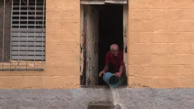 An elderly man pours water out of a bucket from inside his home.