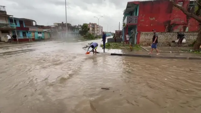 Several people walk along a wide and flooded road, with one scooping a bucket into the flood waters, showing it's quite deep. There are colourful houses in the background.