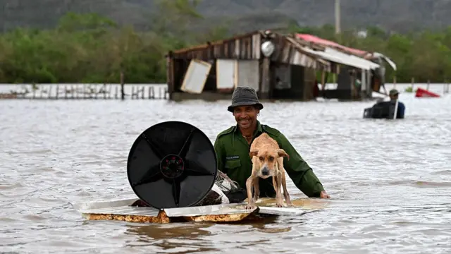 dog on raft pushed through high flood waters by man with flooded house in background