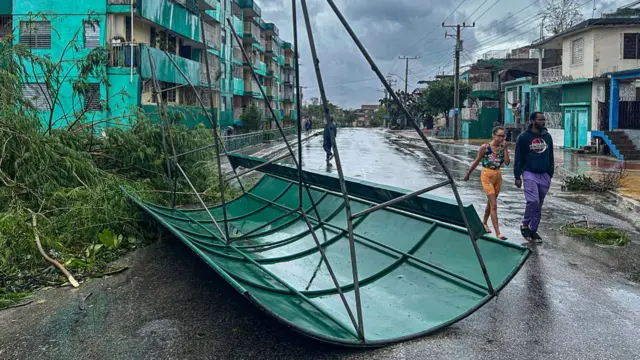 A large piece of green plastic with metal framing lays on a road with people walking beside it