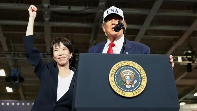 Japanese prime minister Takaichi raises her fist in the air as she stands beside Trump, who is speaking at a podium