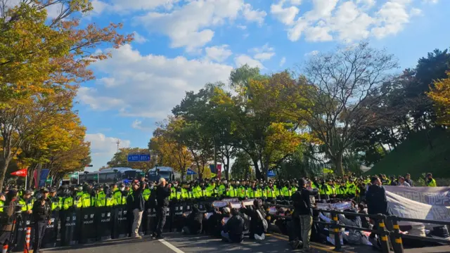 A row of police in neon yellow jackets standing on the road, in front of a group of protesters sitting on the ground