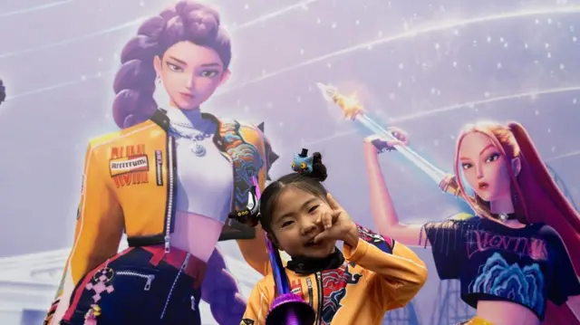 A girl poses in front of a large background with characters from KPop Demon Hunters