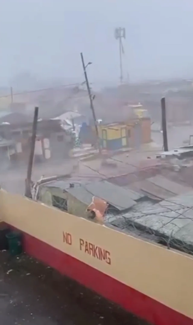 A screenshot of a video which shows strong winds over a city, corrugated roofs are damaged and other debris is visible