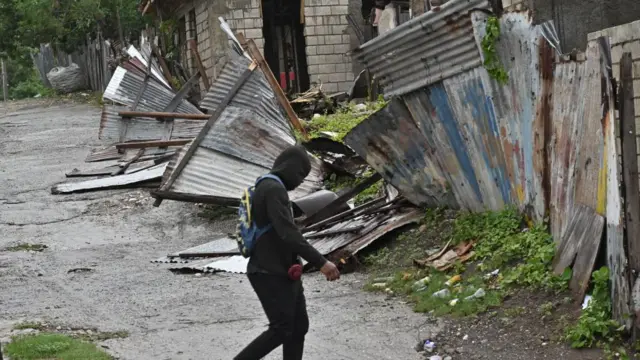 A person walks in front of a house damaged by Hurricane Melissa. Corrugated metal sheets have been blown over and bent in the wind.