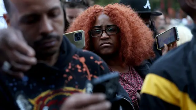 Woman with orange curly hair, black glasses and green eye shadow holds up green phone while frowning, amid other people holding up phones