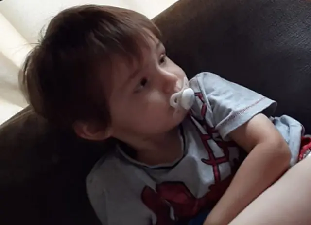 Ethan sitting on a settee with a dummy in his mouth. He has brown hair in bangs and over his ears and is wearing a t shirt with a red cartoon hill and tree
