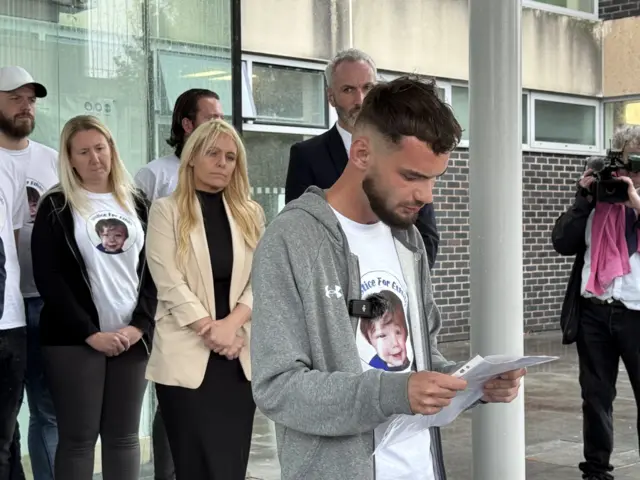 Man in  grey hoodie and white t shirt stood on concrete steps reading from a paper. behind him stands a group of people in smart lcothing looking colemn.