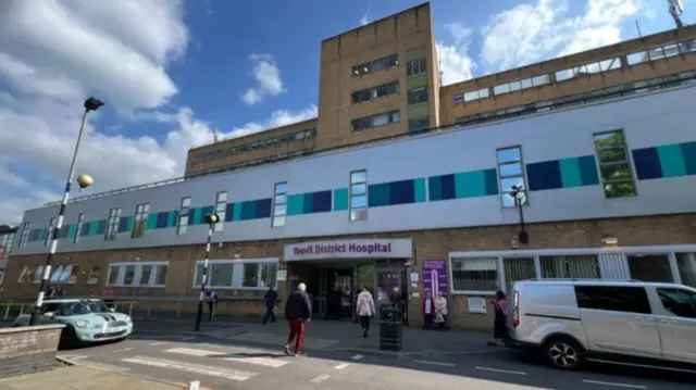 Yeovil District Hospital. It is a brown and white building with blue squares. There are vehicles and people in front of the hospital.