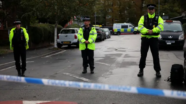 Three police officers wearing hi-vis jackets stand behind a police cordon