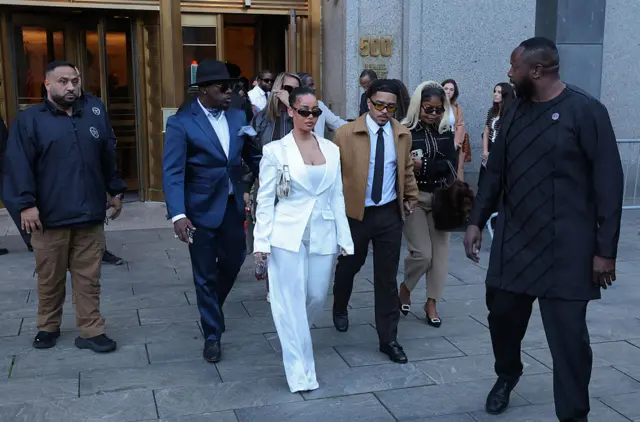 Woman in all white pantsuit and sunglasses walks with man in tan jacket and black pants, also wearing sunglasses