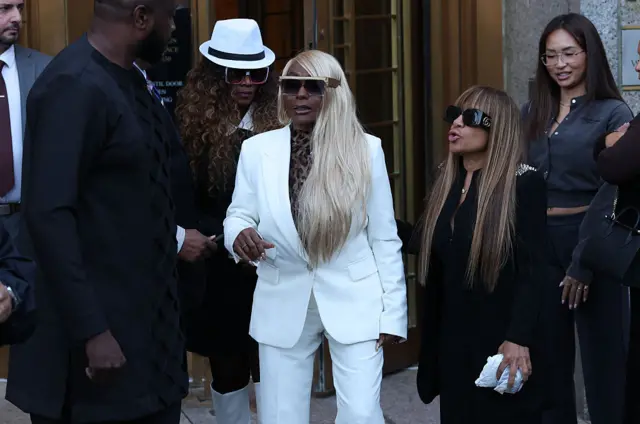 Woman with long blonde hair swept to the side and sunglasses in white suit with leapord-print shirt surrounded by people wearing black and woman in white fedora