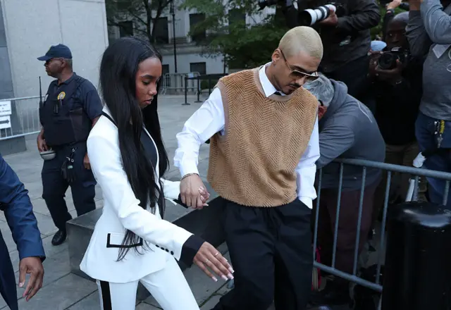 Long-haired woman in white pant suit with black piping holds hand of man with short bleached hair in tan sweater vest, white shirt, black pants
