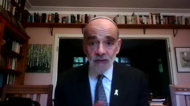 Rabbi Wittenberg is sitting in a room with a pink wall and many books behind him.