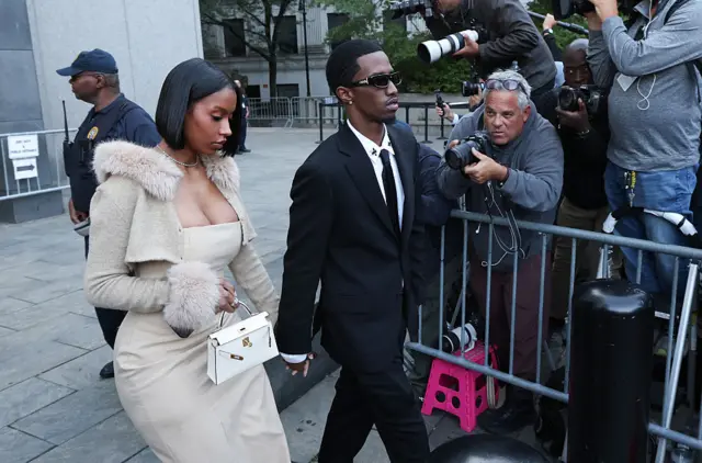 Woman in tan low-cut dress with tan fur-trimmed bolero and small white handbag holds hands with man in black suit wearing sunglasses as they pass photographers