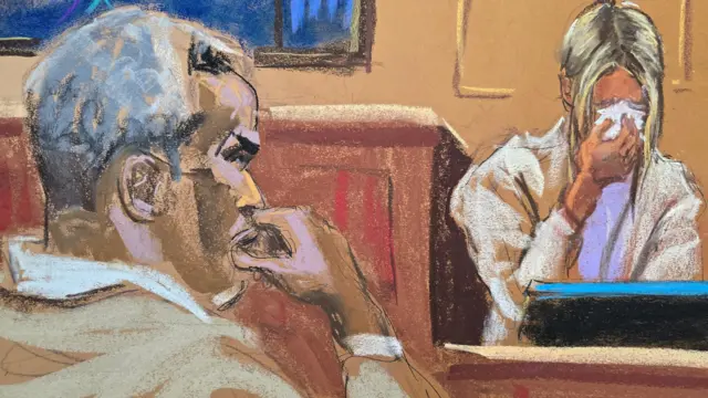 Combs is in the left side of the picture, hand to his mouth, wearing a tan suit with white hair, Mia is in the right side in the witness box with blonde hair hanging down and blotting her tears with a tissue, obscuring her face