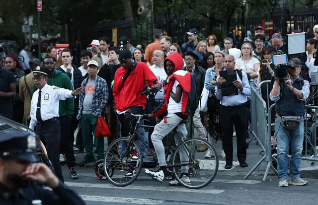 Group of people clustered behind traffic cop in white shirt with hand out, with variety of expressions from bored to interested, and a cameraman next to security fending
