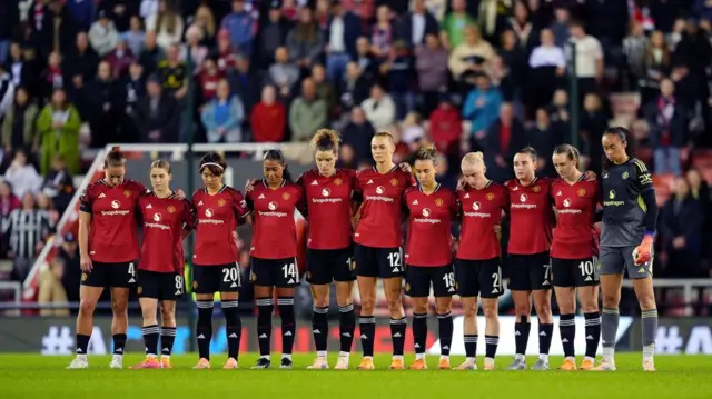 A women's football team wearing red shirts, black shorts and socks, stood in a line in front of packed stands holding one another. A few look up, the rest look down solemnly