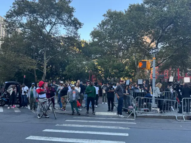 Line of people on sidewalk and in striped crosswalk, some behind security fencing, with green trees behind them