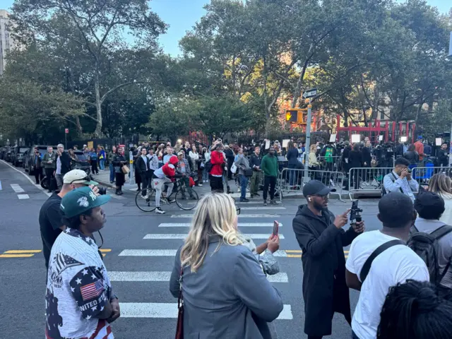 Crowds of people on both sides of the street with a striped crosswalk between them, many holding up phones and cameras
