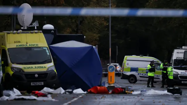Police and ambulance vehicles can be seen parked near a blue tent. Some white sheets lie on the ground. Three police officers can be seen on the right hand side