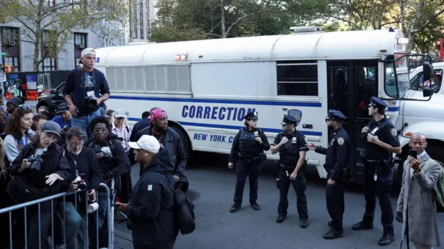 Side of white bus with caged windows labeled "CORRECTION" in blue, with four uniformed police in front and photographers huddled behind security gate to side