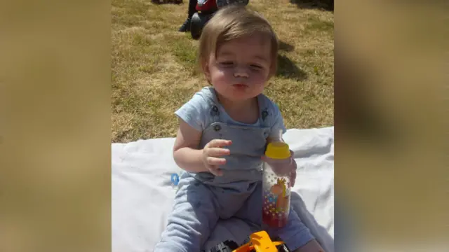 Ethan is sitting on a blanket outsiside on some grass, He has on coverall and a blue t shirt and his hold a juice bottle