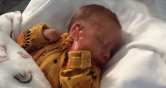 A newborn baby wearing a yellow babygrow. He has his hand by his face and is under a white blanket with rainbows on it.
