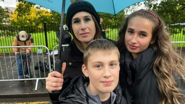 A family of three, a mother and a son and a daughter, stand in a park under a blue umbrella
