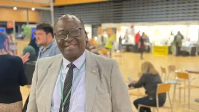 A picture showing Mary-Odoy smiling at the camera standing  a hall with some people stood and one sat at a table behind him