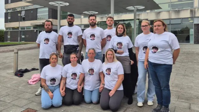 11 members of Ethan's family stand and knee outside court wearing t-shirts with his face on.