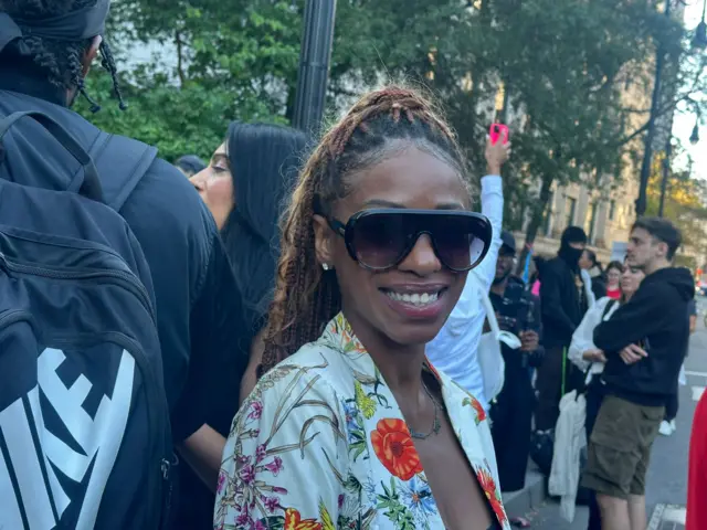 Woman in sunglasses and white jacket printed with flowers smiles at the camera