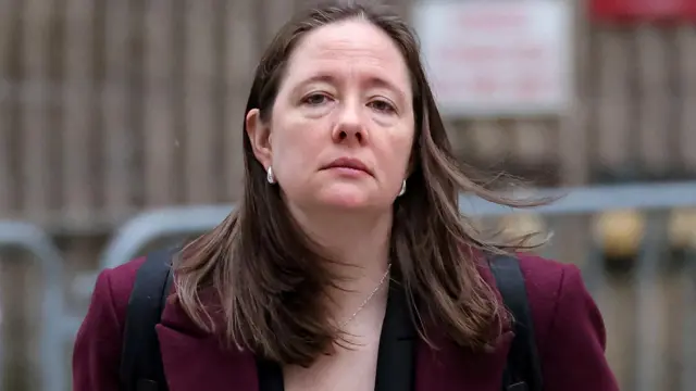 Maureen Comey with breeze lifting her hair, wearing silver hoop earrings, necklace and maroon blazer