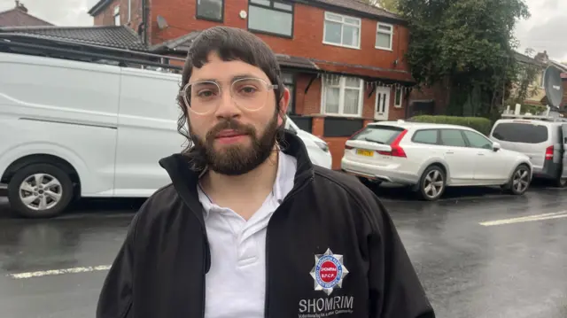 David Elkouby, who is an Orthodox Jew, stands in the middle of a road on a wet day wearing a black jacket and white polo shirt.
