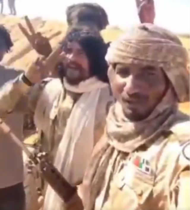 Abu Lulu wearing camouflage clothing and holding an assault rifle and putting up a v for victory sign with his index and middle finger, standing next to and another RSF member