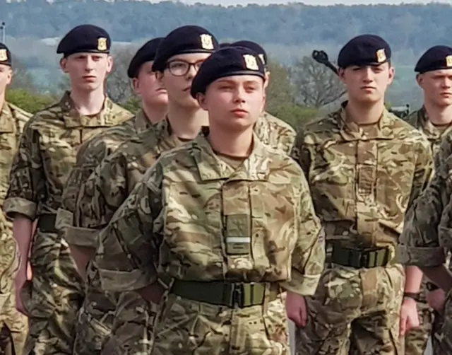 Gunner Beck wearing her camouflage military uniform and black cap. She is standing among her fellow soldiers with her arms clasped behind her back and a stern facial expression.