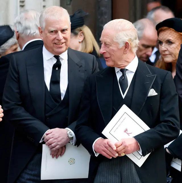 Prince Andrew and King Charles III stood next to each other in black suits, their hands are crossed.