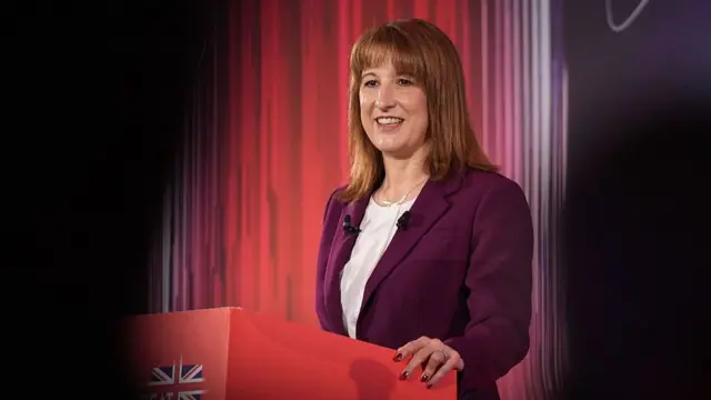Mid-shot of Rachel Reeves in deep purple blazer, white shirt as she leans on red podium