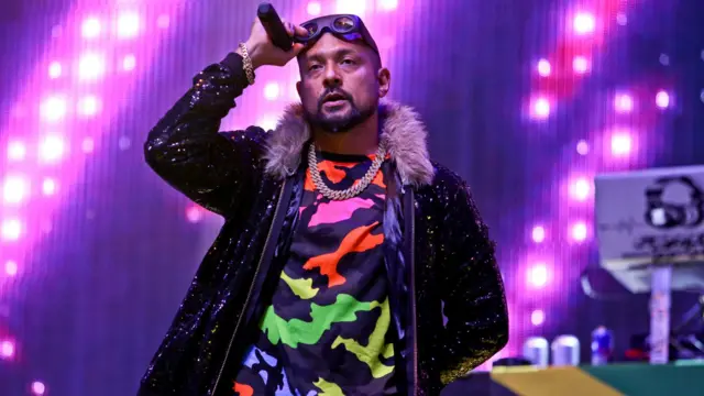Sean Paul wears a colourful camouflage shirt and sparkly jacket while performing on stage
