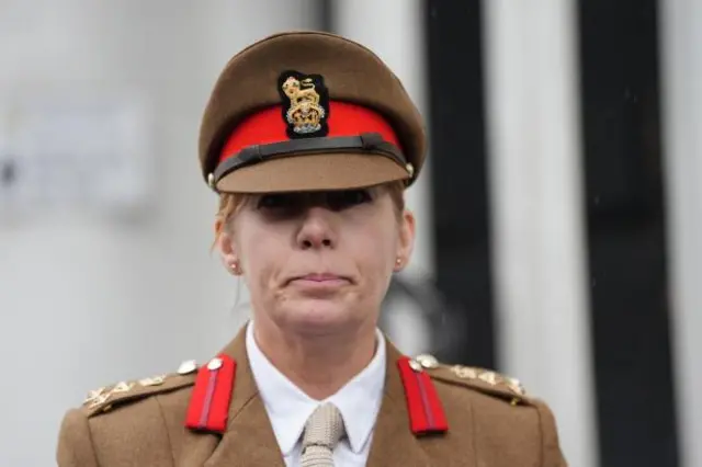 Brigadier Melissa Emmett wearing a brown military suit with gold gilded epaulettes and a matching cap.